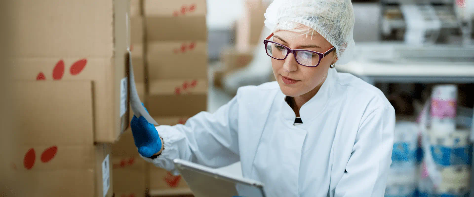 A worker in sanitary outfit looks at a tablet while standing next to stacked boxes A worker in sanitary outfit looks at a tablet while standing next to stacked boxes