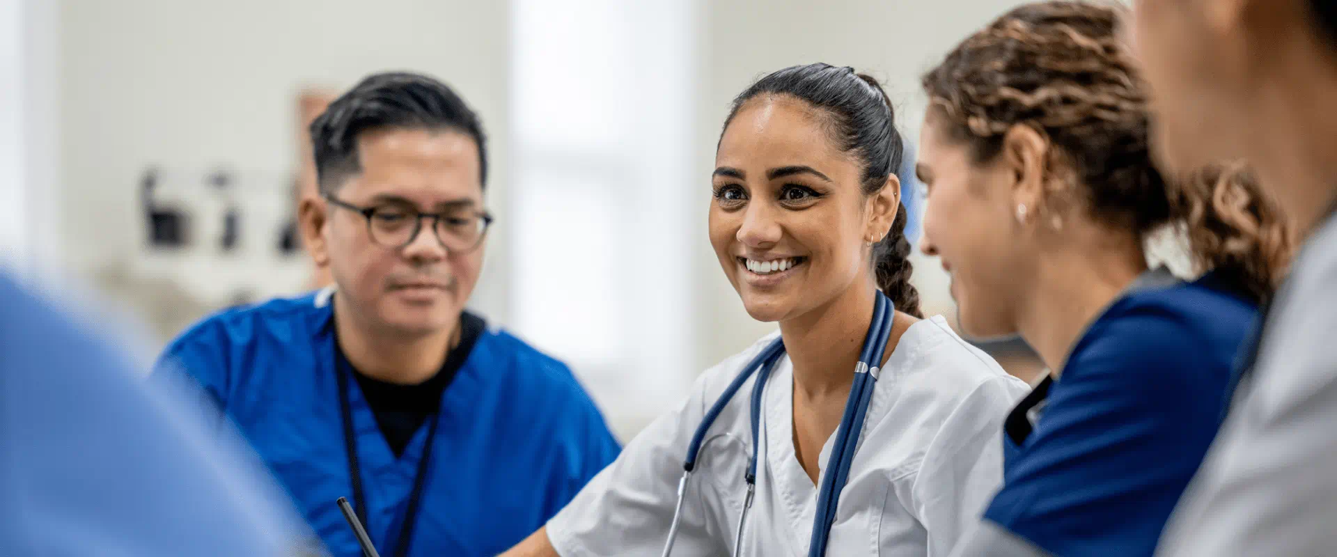 A group of nurses and healthcare workers in scrubs stand talking and smiling at a nurses station.