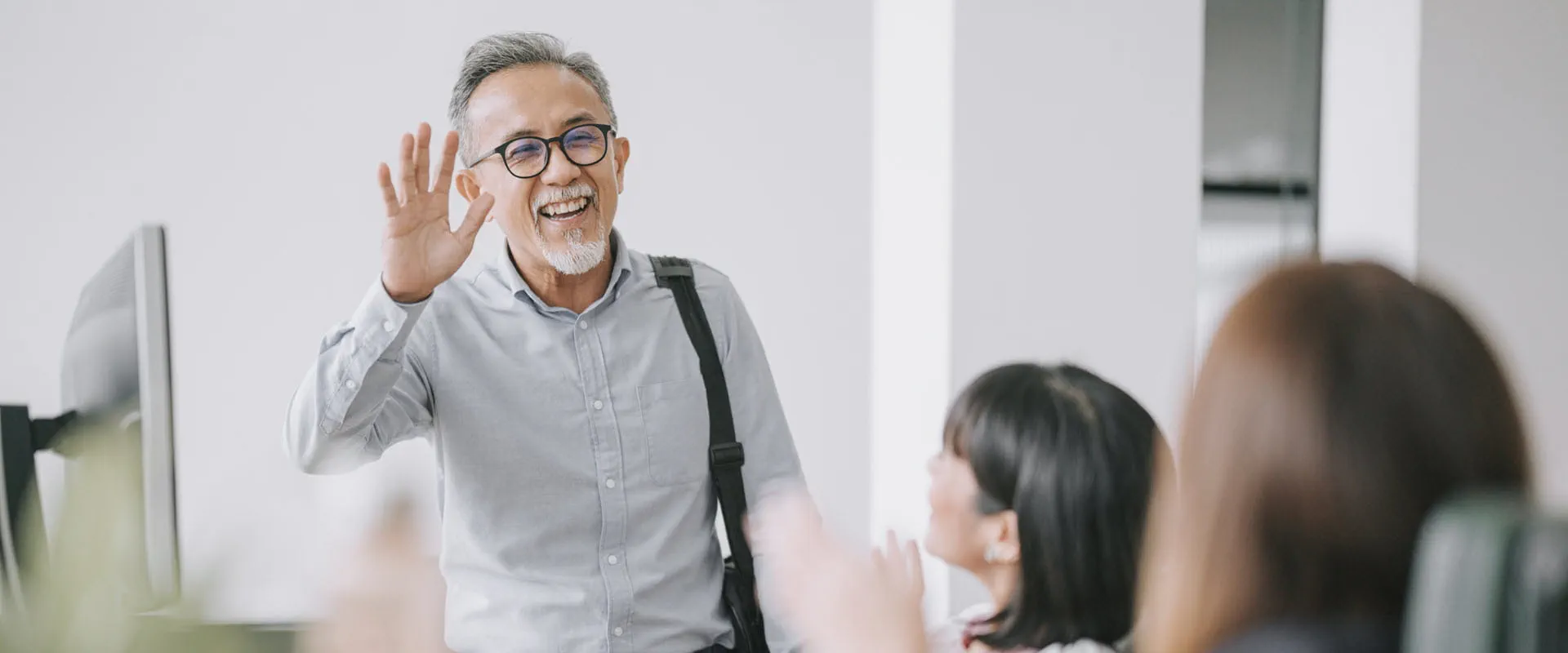 A smiling person with glasses and a gray shirt waves as they enter a modern office space. 