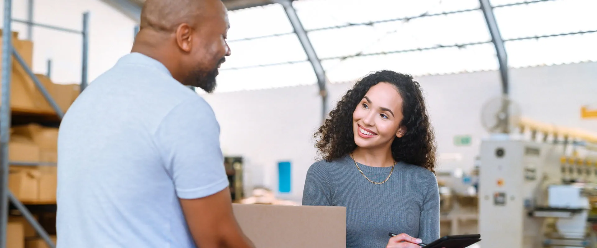 Two workers talk in a shipping facility. One is carrying a box, the other is holding a tablet.