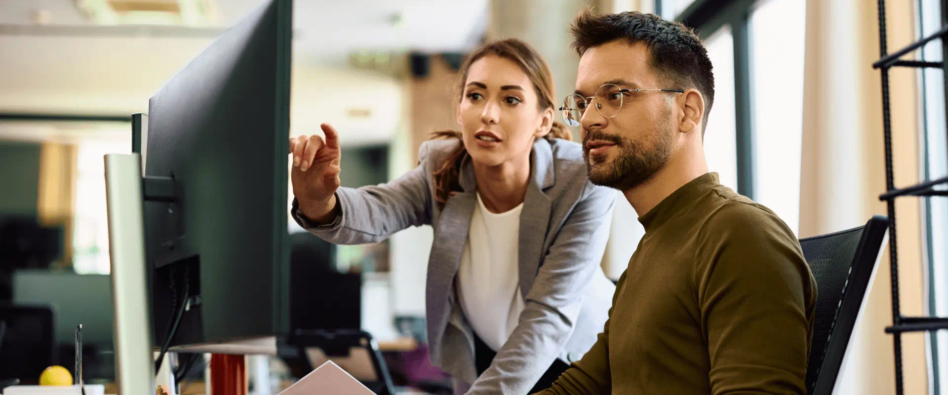 Two people look at a computer screen. One is sitting at the computer, the other is standing next to them, pointing at the screen. Two people look at a computer screen. One is sitting at the computer, the other is standing next to them, pointing at the screen.