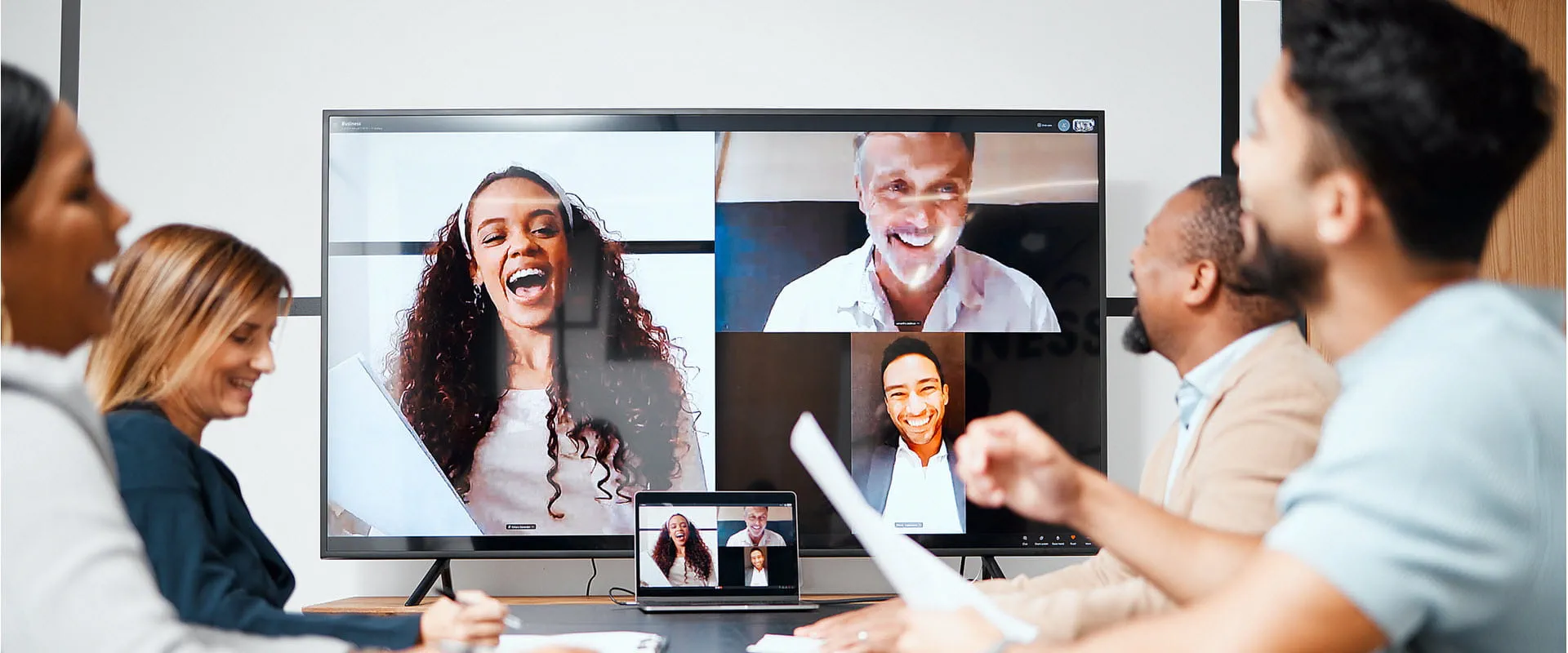 A small group of business people sit and laugh in a conference room. They are speaking to three coworkers on a large monitor, who are video-calling in to the meeting.