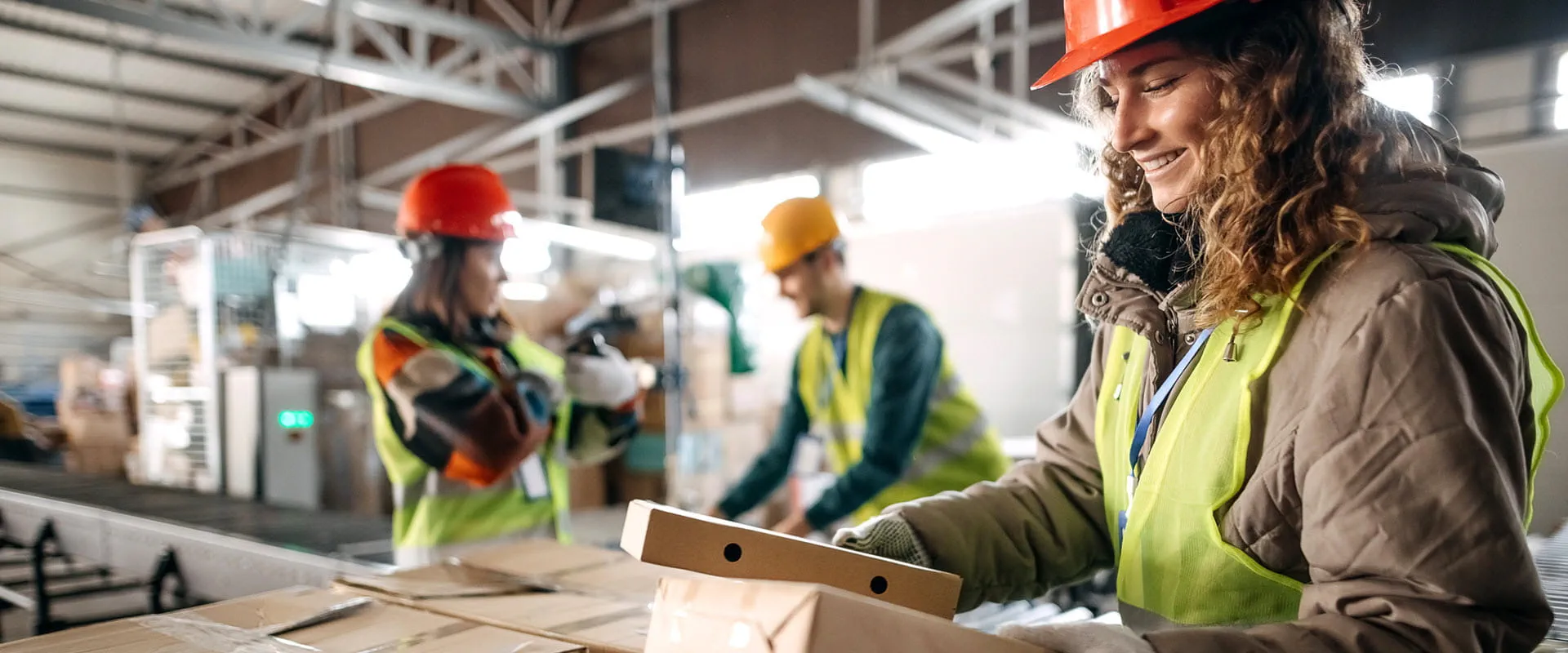 Three workers in a distribution center wearing hard hats and safety vests, sort packages.