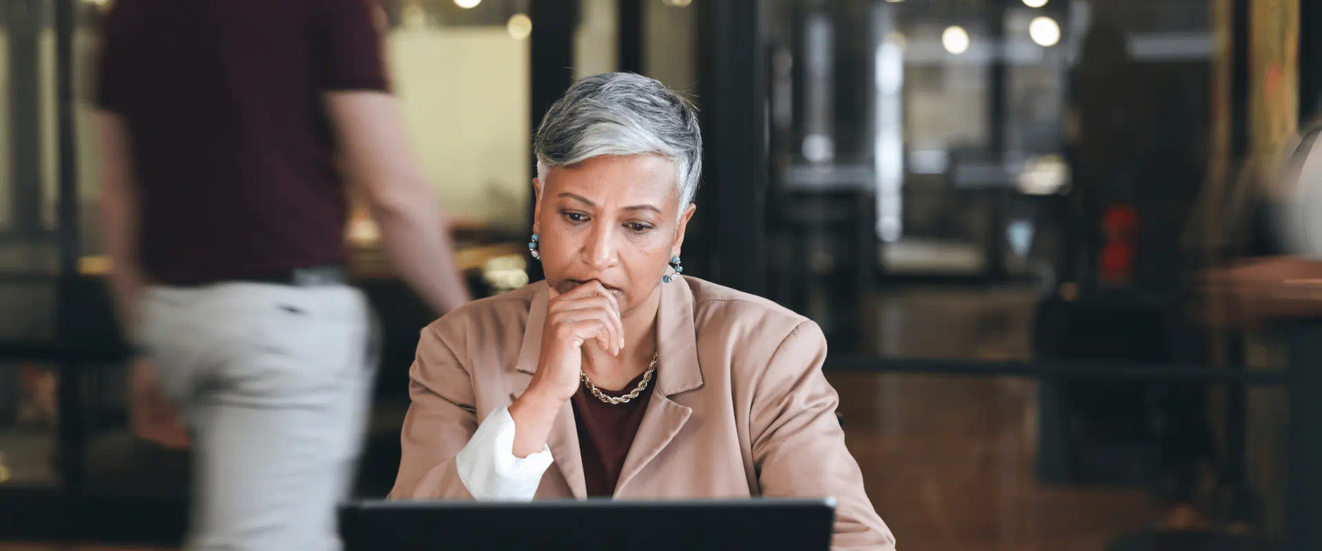 A manager sits at a laptop looking stressed, someone is walking out of the office behind them.