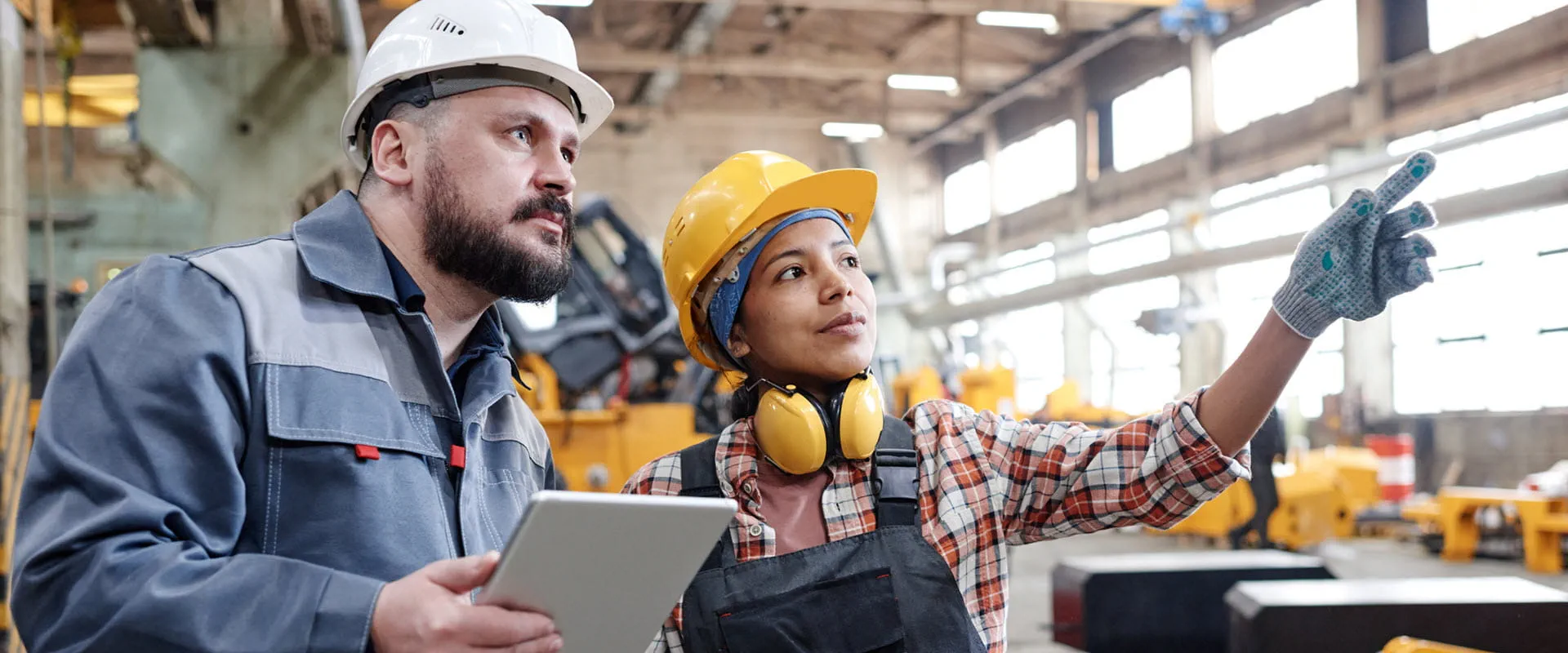 Two workers in hard hats and coveralls look at something in the distance in a manufacturing warehouse.