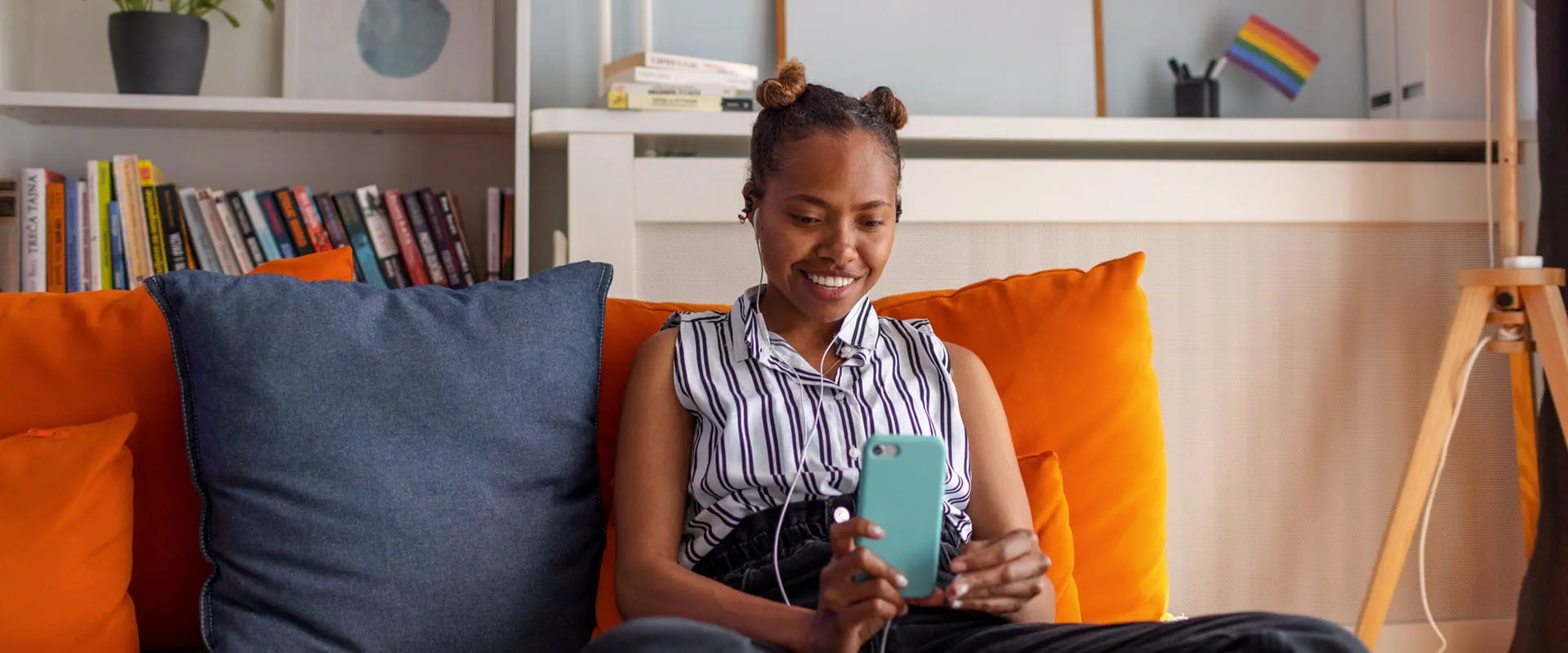 A fashionably dressed young person sits on an orange couch looking at their phone with headphones in