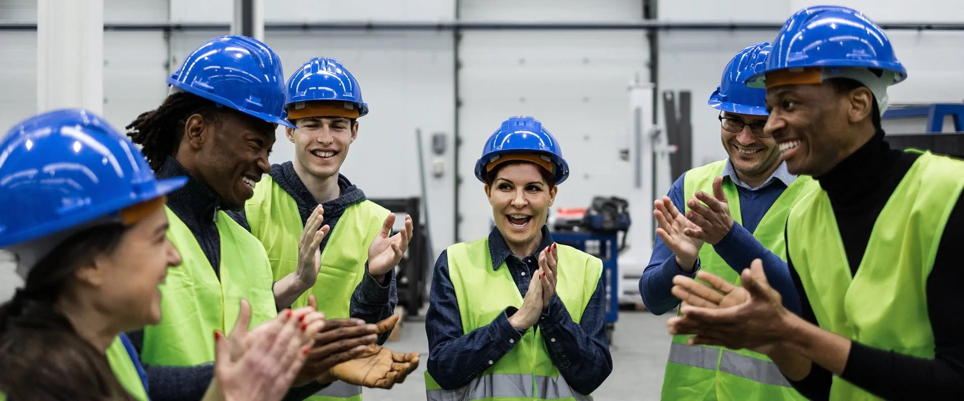 A group of workers in hard hats and safety vests, stand in a semi-circle, clapping and smiling