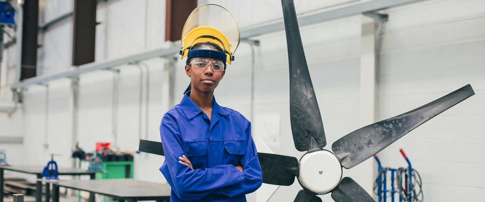 A woman in blue coveralls, safety glasses, and a hard hat, stands in front of an unfinished windmill propeller, in a workspace