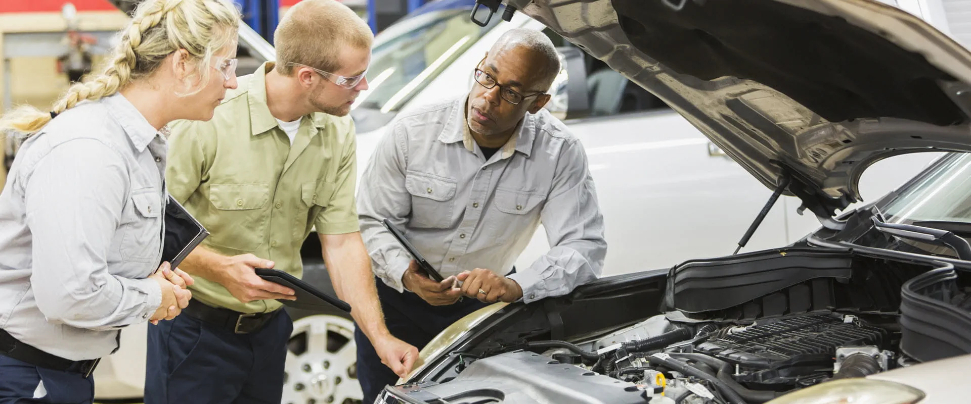 Two younger workers stand next to an older worker, who is explaining something about the car in front of them, which has its hood open.