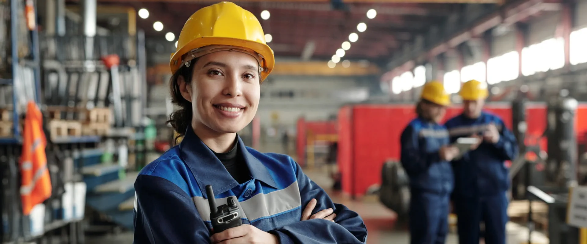 Young smiling worker of modern industrial plant or factory in workwear and protective helmet standing in large workshop.