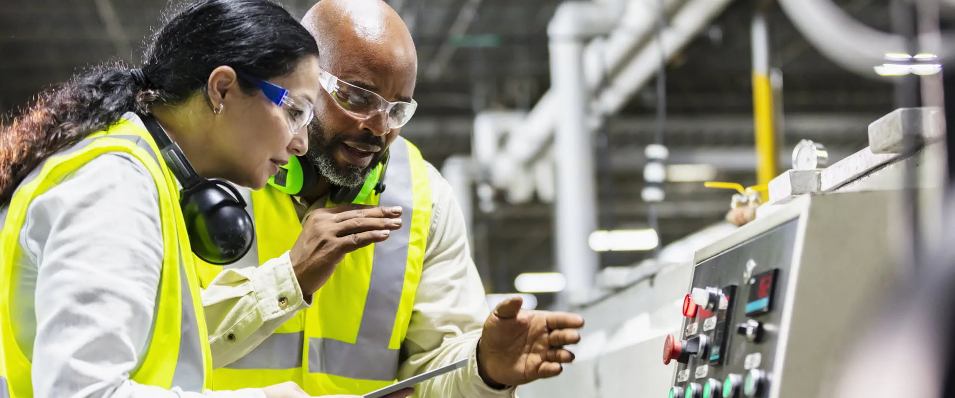 Two workers are in yellow safety vests and safety glasses. One worker is explaining something to the other while they look at a piece of equipment.