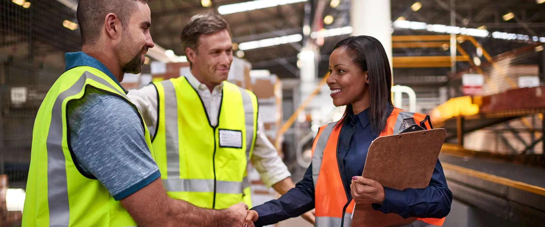 Three workers in business casual dress and safety vests, stand on a warehouse floor having a meeting.