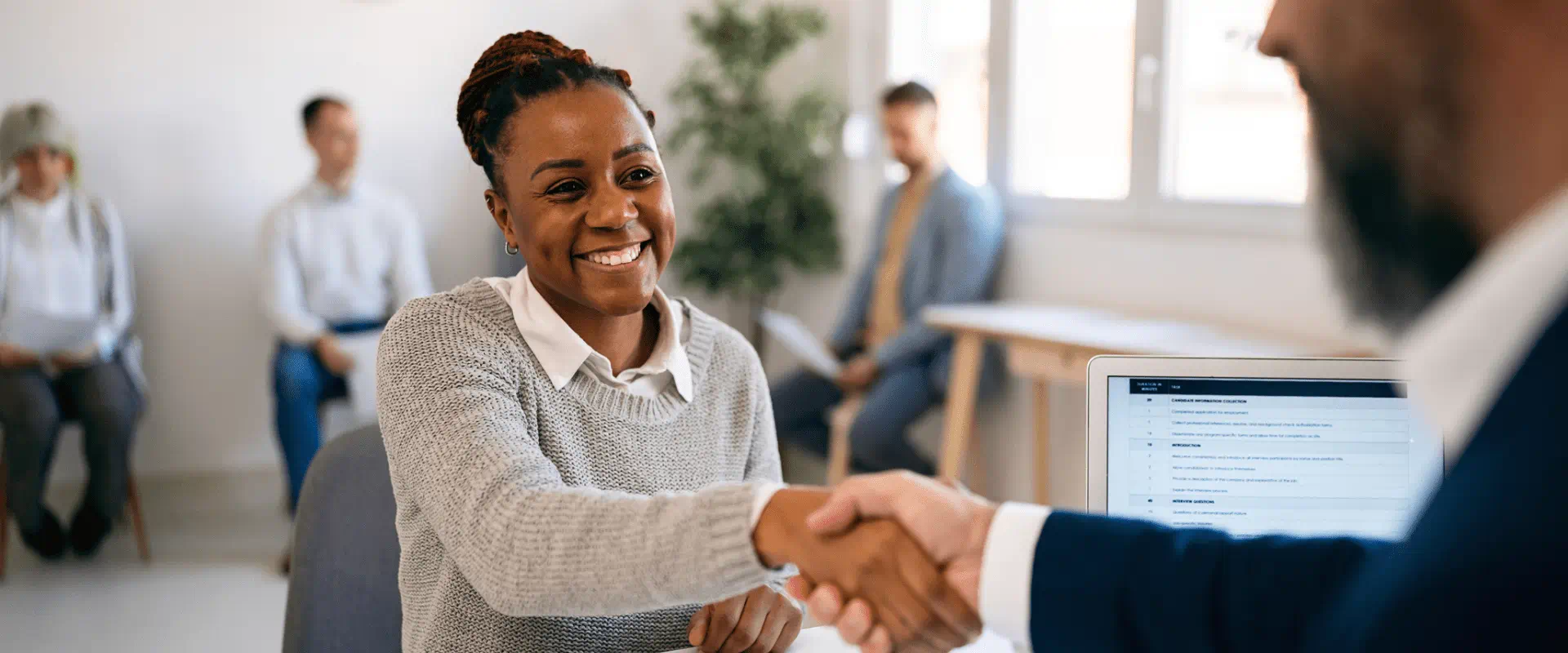 A smiling job candidate shakes hand with an interviewer. Behind them, others sit at chairs waiting to be interviewed.