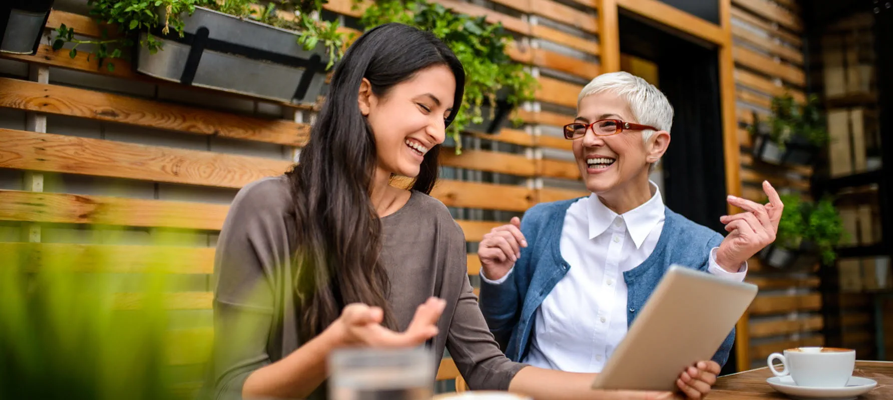A younger and older coworker sit next to one another at a coffee shop. They are laughing.