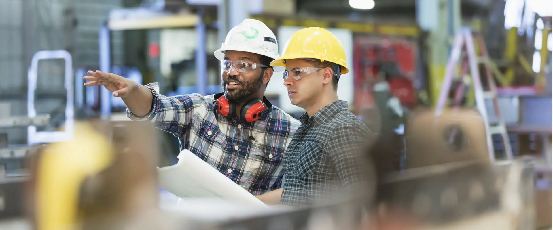Two industrial workers wearing hard hats and safety glasses stand in a manufacturing facility. 