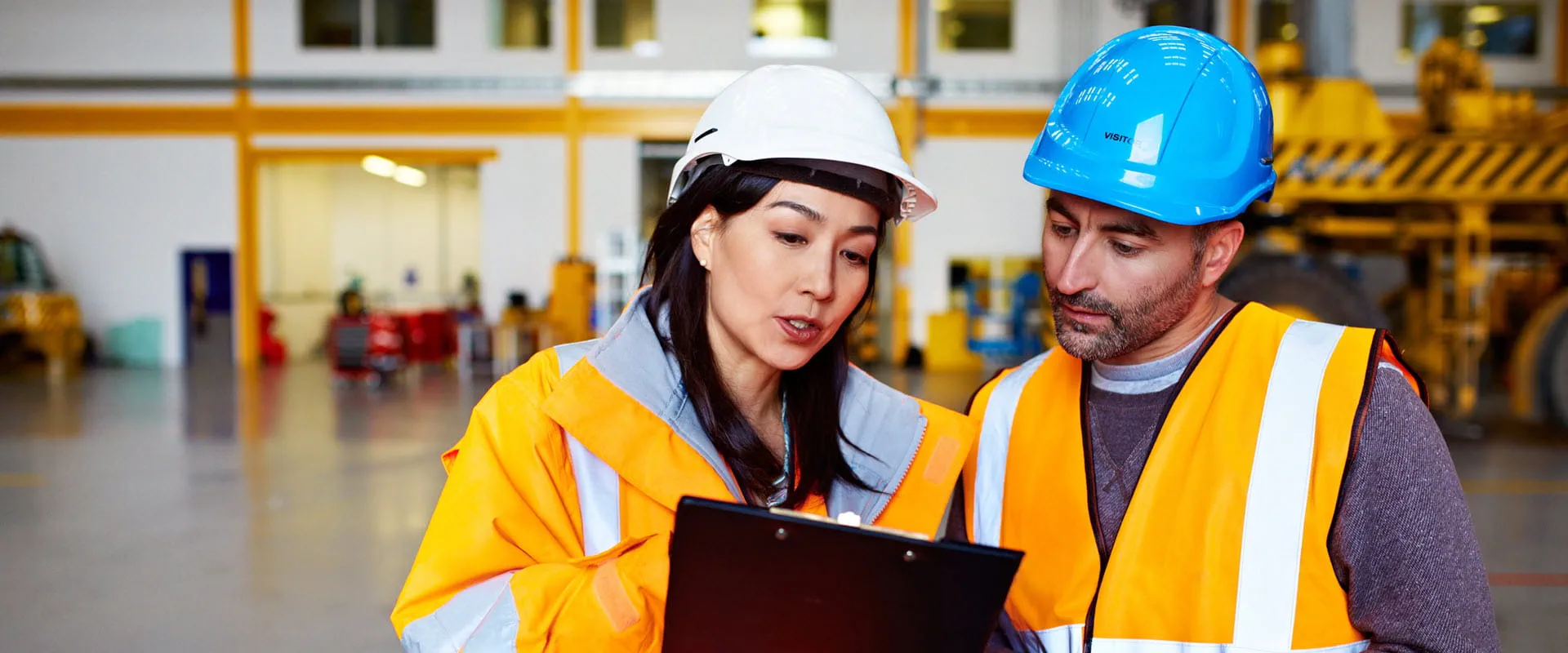 Two workers wearing hard hats and safety vests stand in an empty warehouse discussing something in the distance