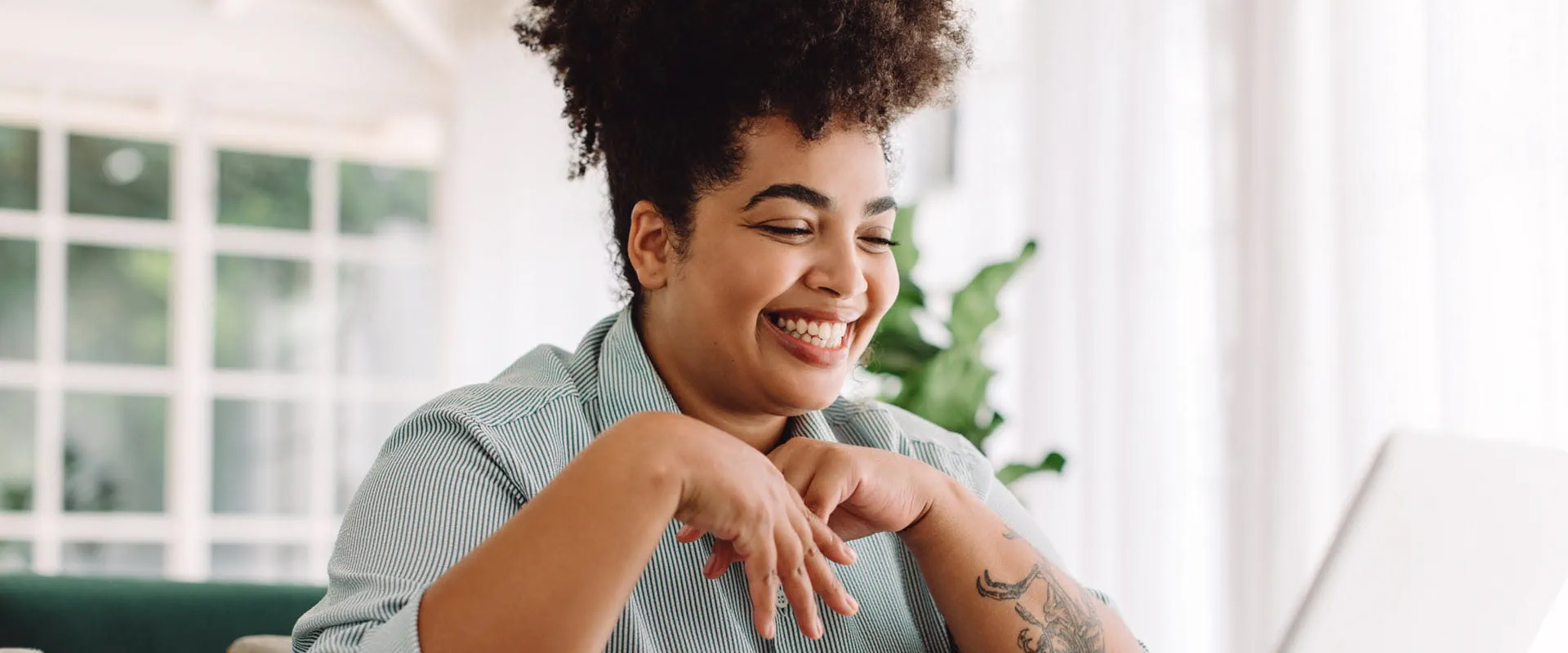 A smiling individual with curly hair and tattoos works on a laptop in a bright, home-like setting.
