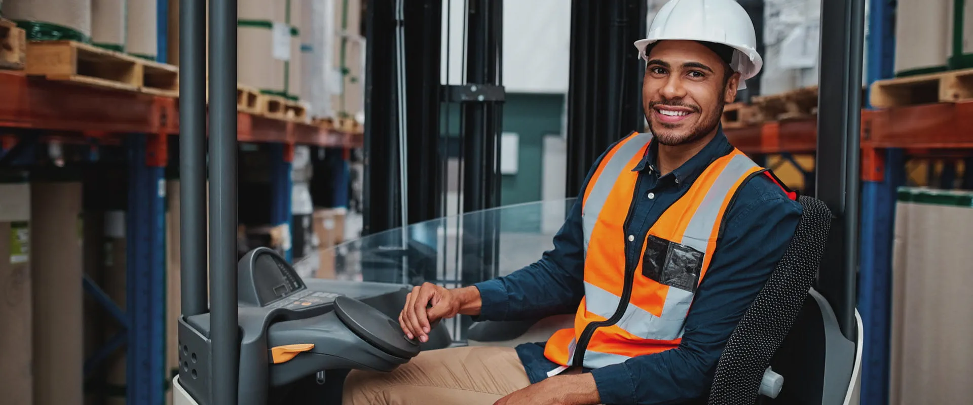 Forklift operator smiling, looking at camera, wearing hard hat and orange safety vest.