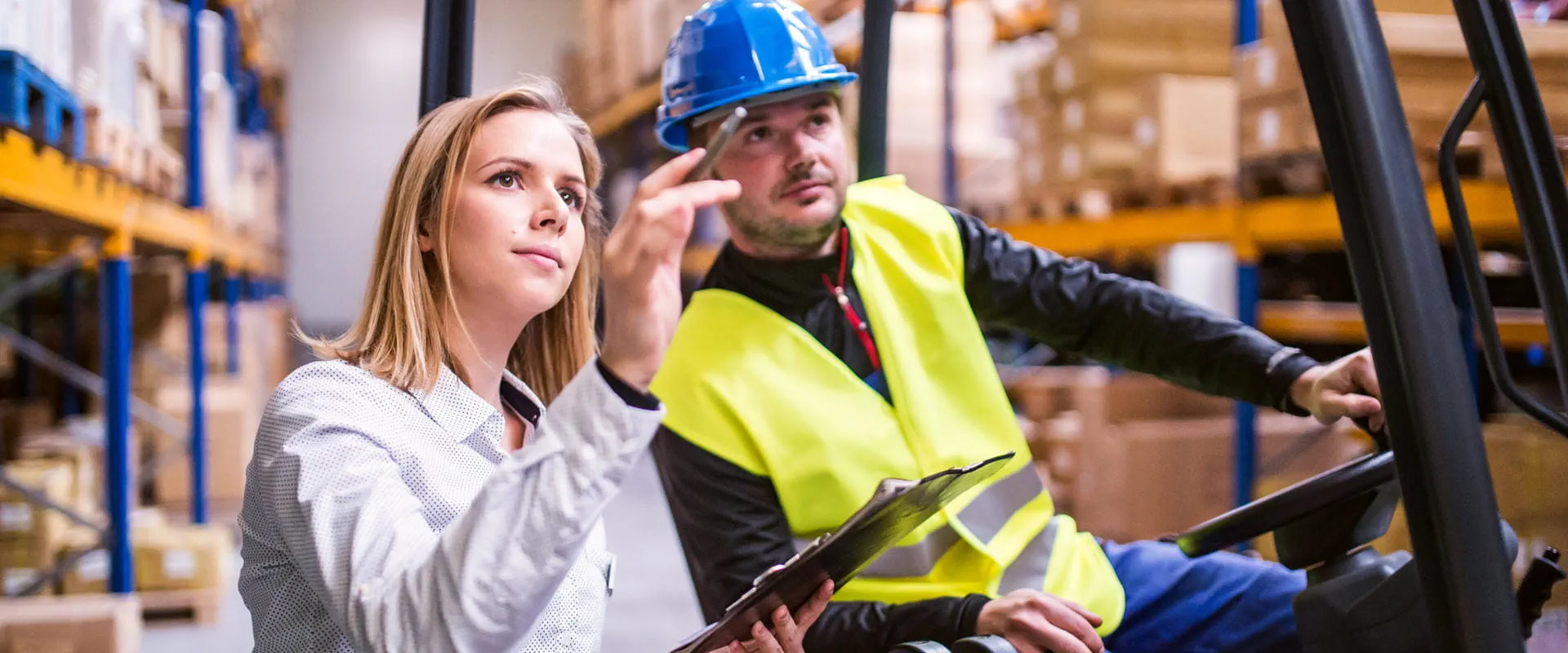 A forklift operator in a warehouse speaks with a manger in a suit, who is pointing at something across the floor
