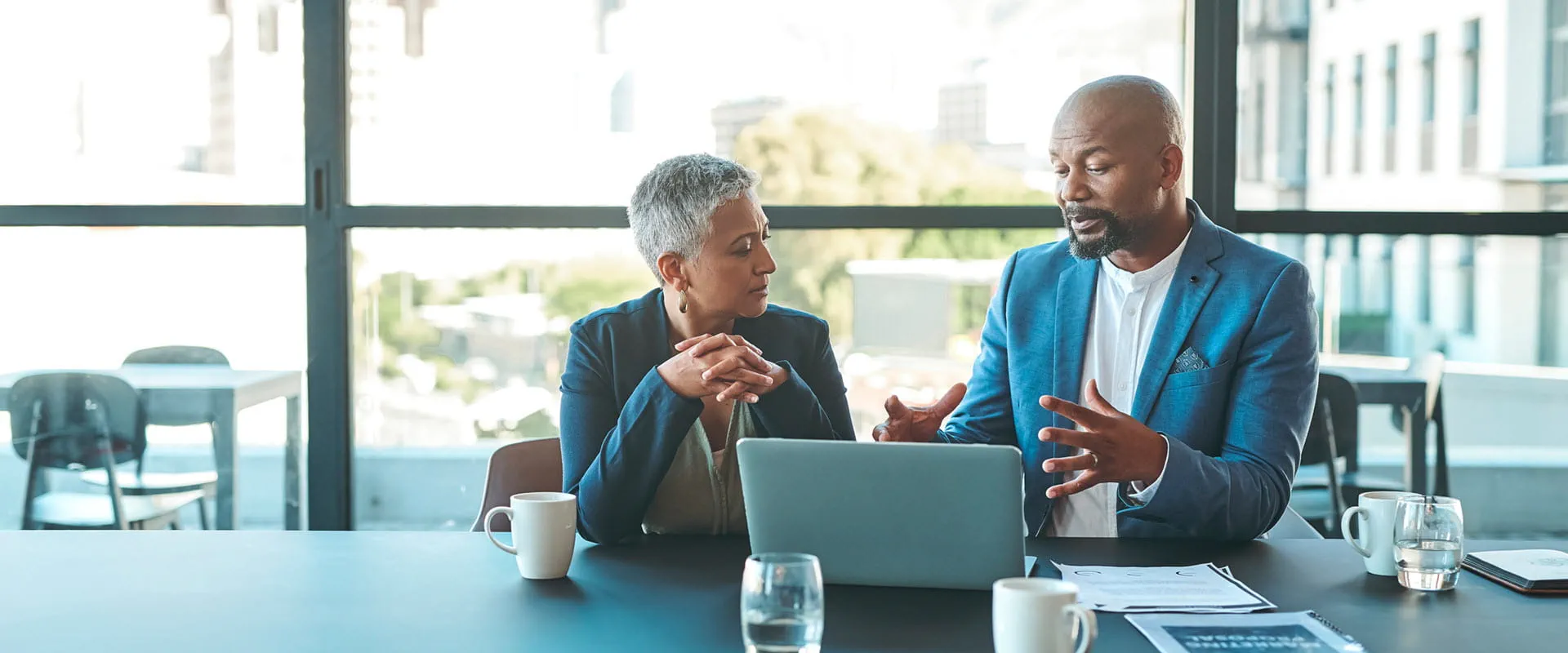 Two business professionals having a discussion at a table in a modern office, with a laptop and documents in front of them.