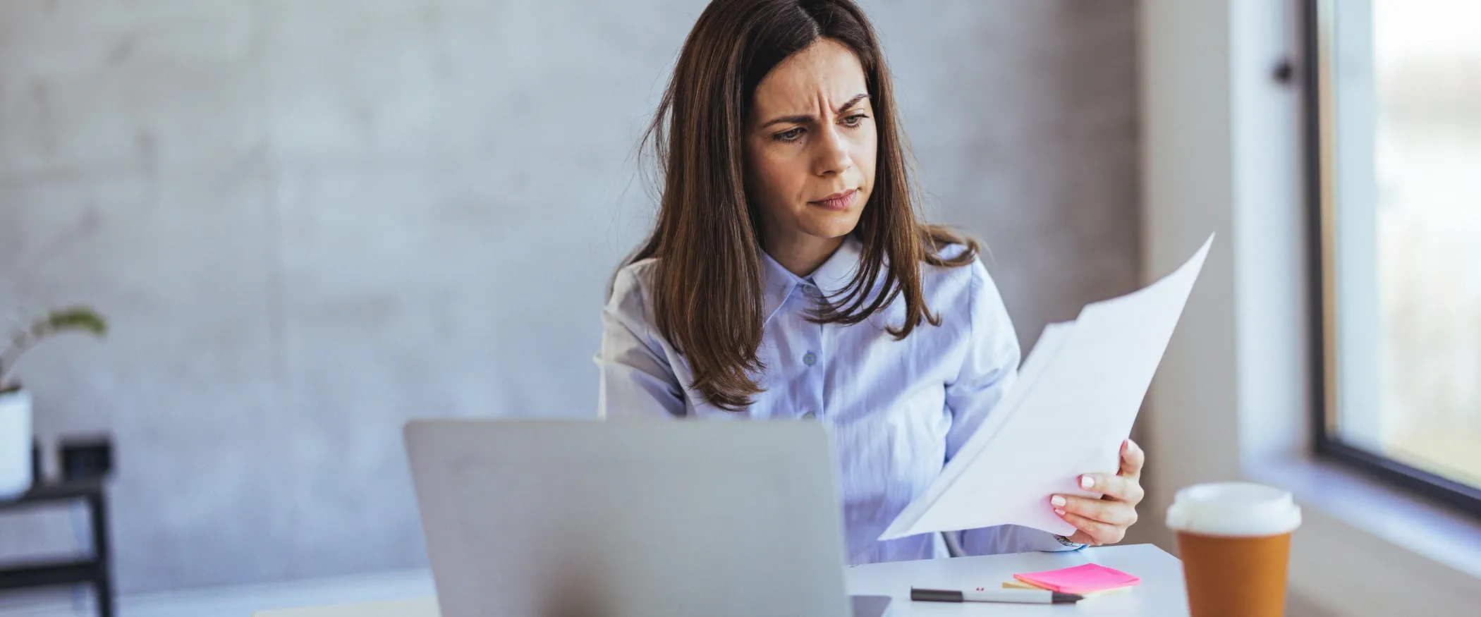 A manager sits at a desk looking at sheets of paper, with a laptop and coffee cup on the table
