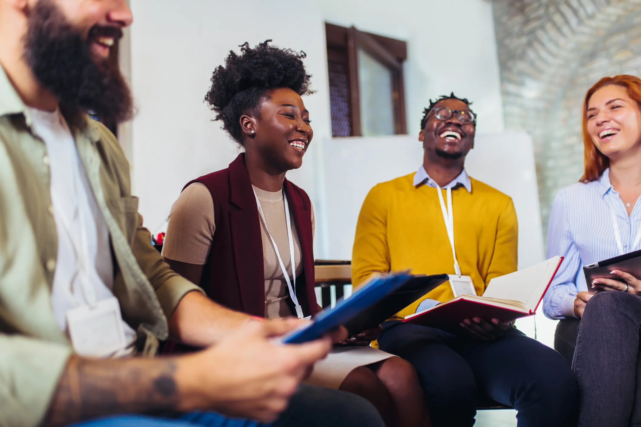 Group of diverse professionals laughing and engaging in a collaborative discussion during a team meeting, holding notebooks and tablets.