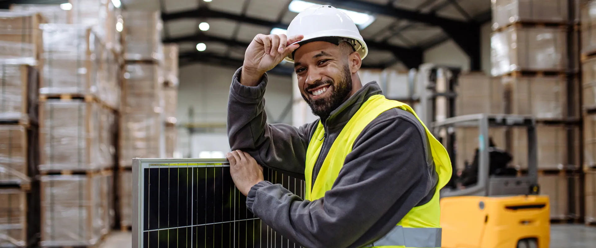 Smiling worker wearing a hard hat holding solar panel in warehouse.