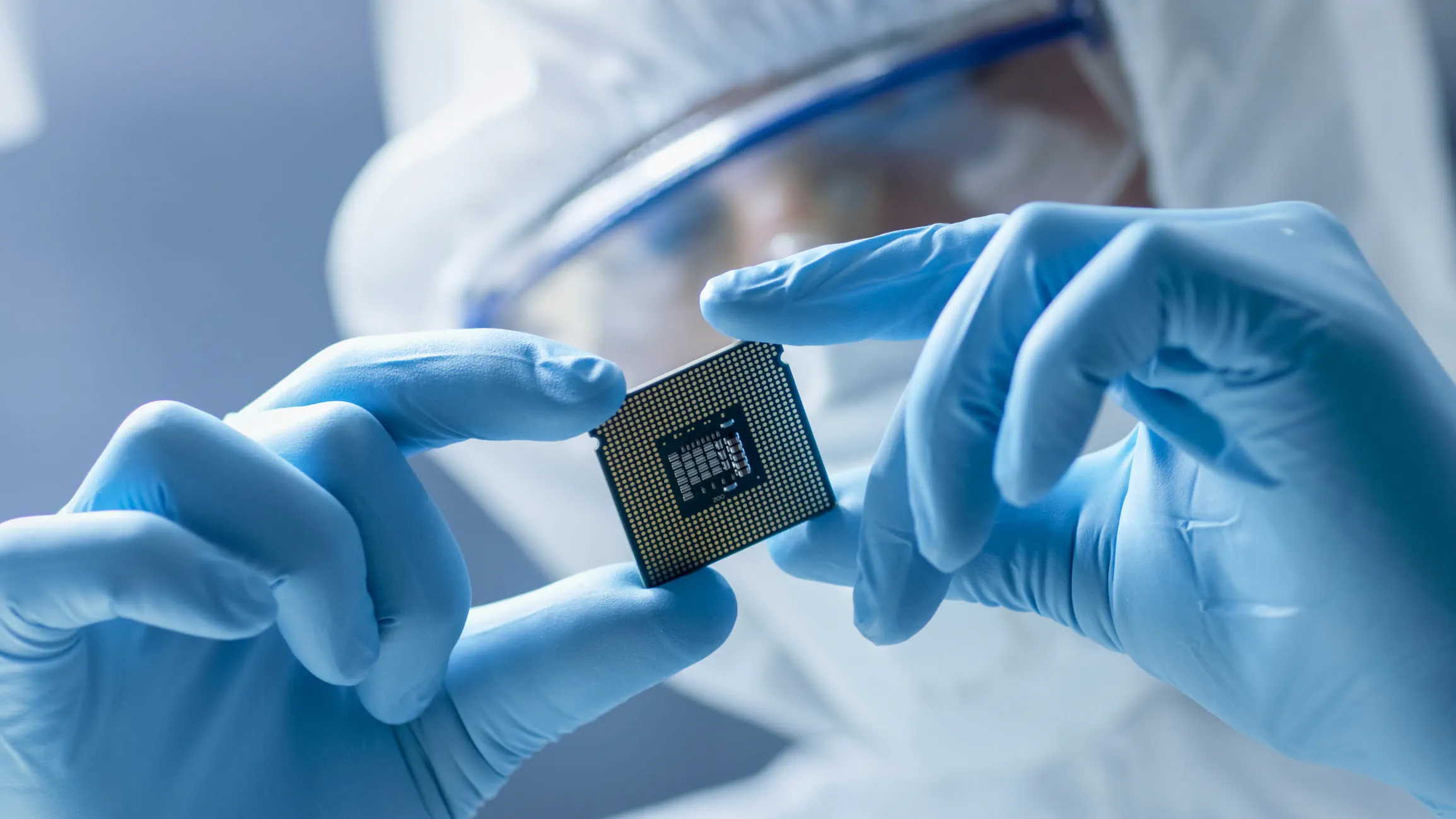 A technician wearing blue gloves and protective gear carefully holding a microchip in a cleanroom environment.