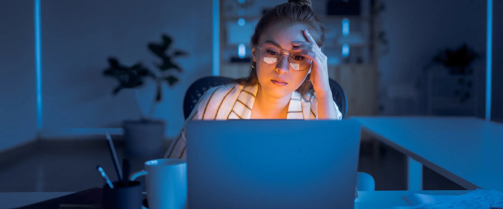 A young worker sits alone in a dark office. They seem stressed and are focused on their laptop screen.