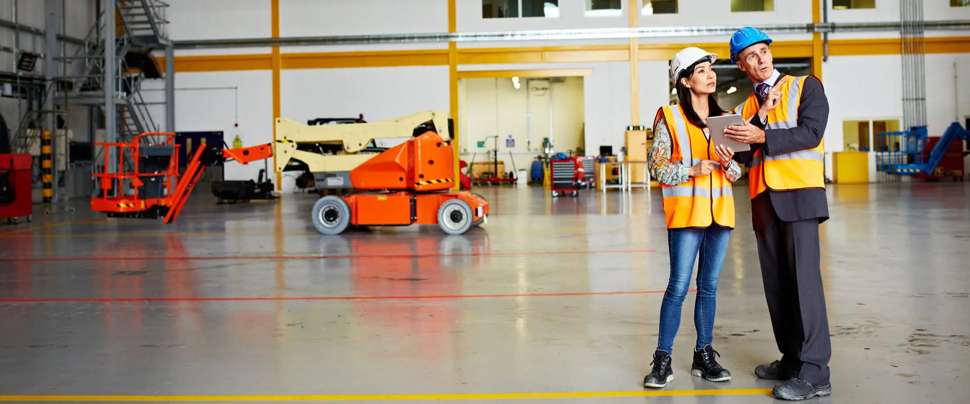 An individual, in a suit, hard hat, and safety vest, shows their tablet to another person, in jeans, a hard hat, and safety vest, in an empty warehouse