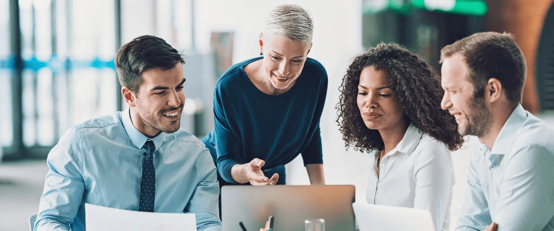 Four workers in business clothes sit around a laptop at a conference table. One of them is pointing at the screen.