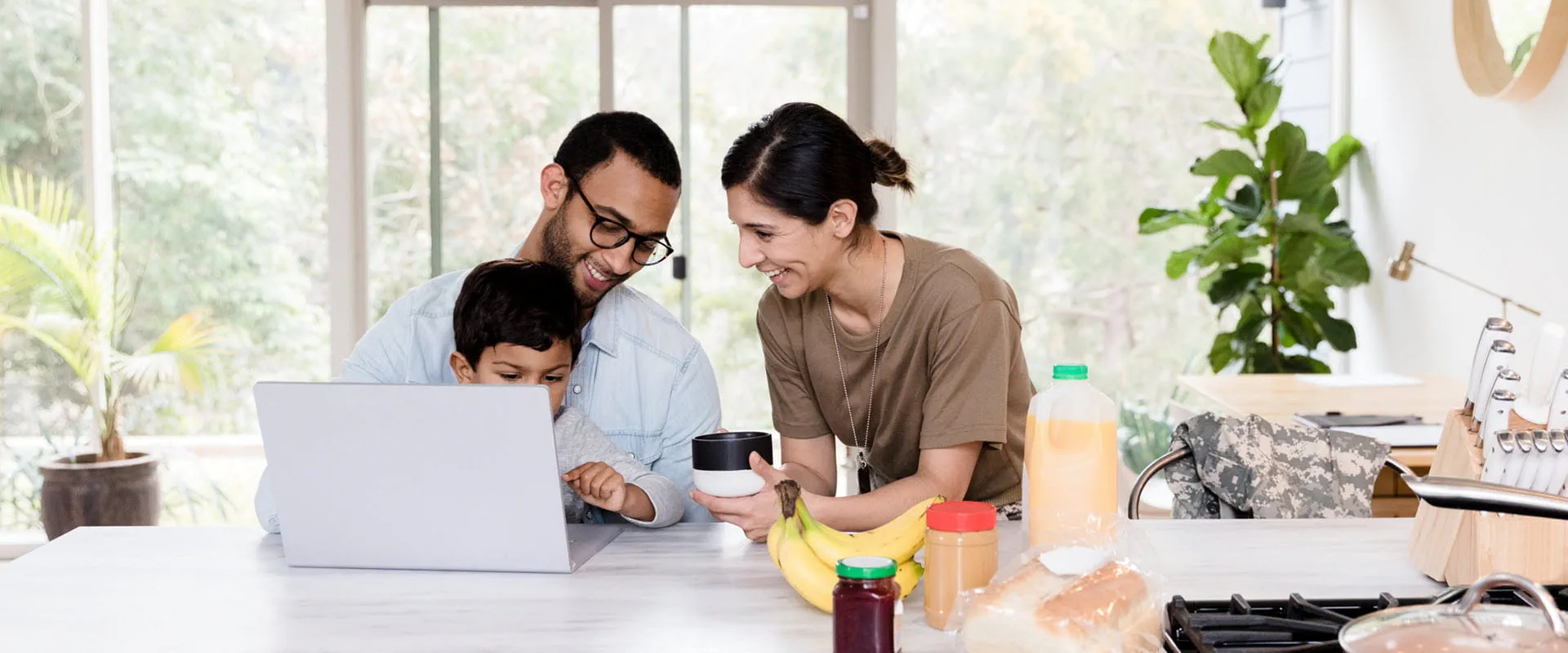 Two parents, one in a military uniform, sit at a kitchen island with their child in between them. There is a laptop and breakfast on the table.
