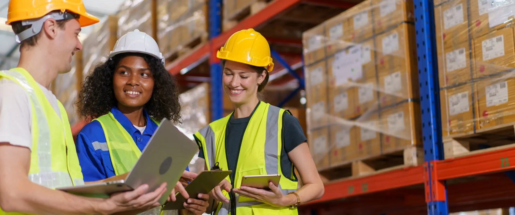 Three workers in hard hats and safety vests stand in a distribution floor. They are talking.