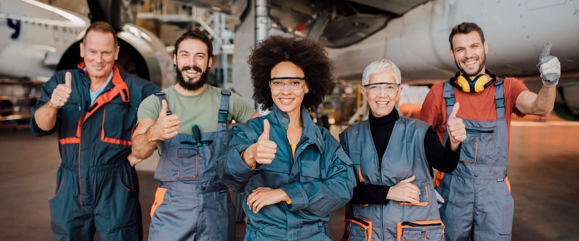 Group of diverse aviation maintenance professionals smiling and giving a thumbs-up while standing in front of an aircraft in a hangar. Group of diverse aviation maintenance professionals smiling and giving a thumbs-up while standing in front of an aircraft in a hangar.