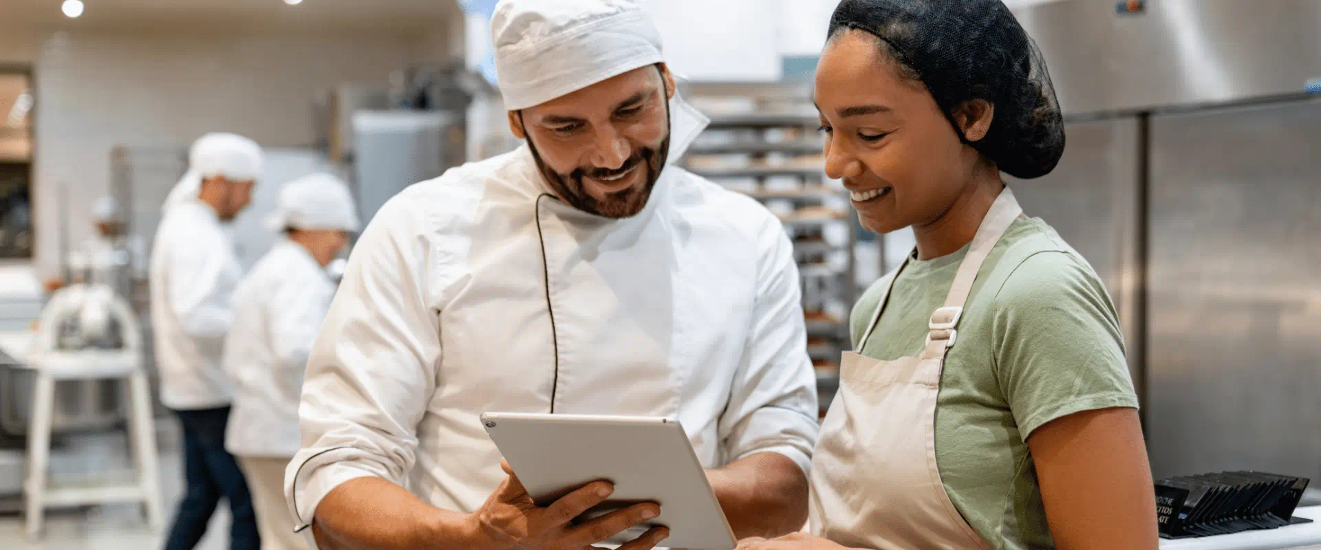 Two workers in hair nets and aprons talk to each other while looking at a tablet. They are in a commercial kitchen.