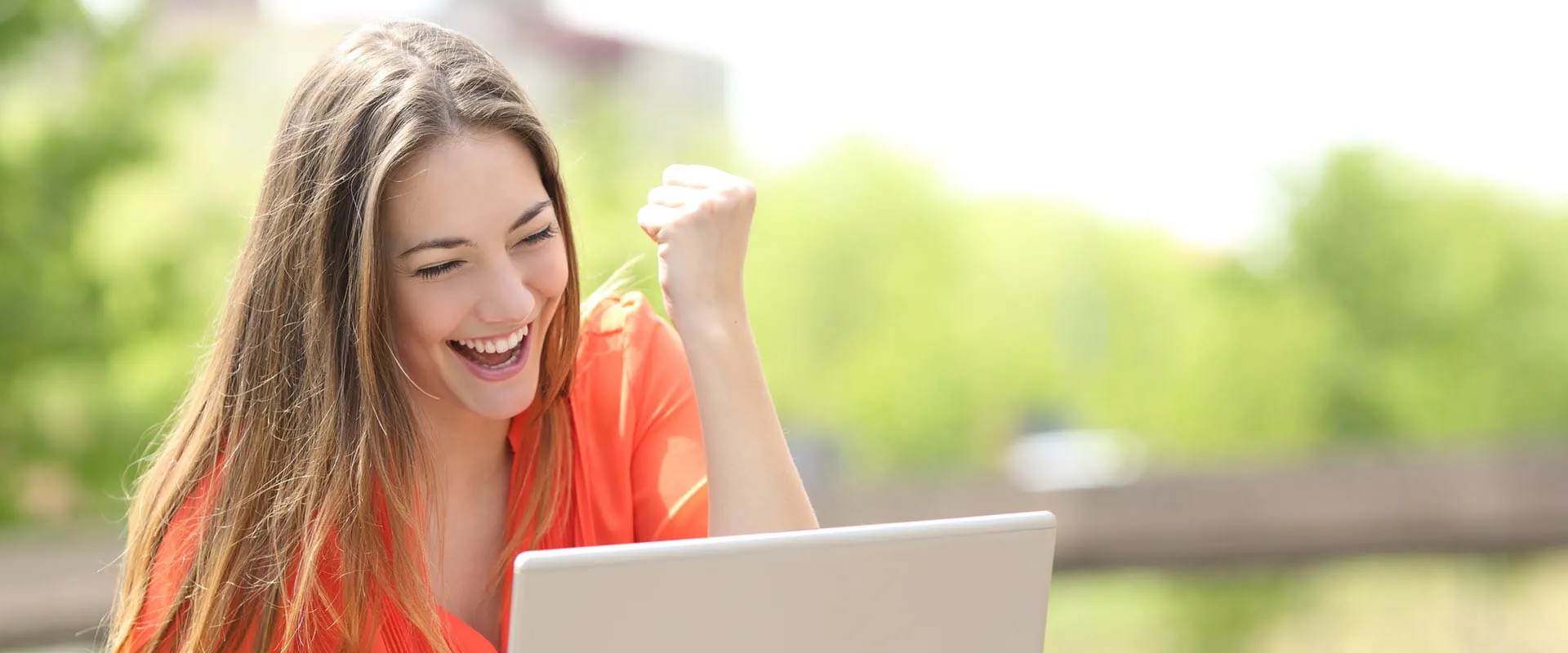 A person sitting outdoors, wearing an orange blouse, smiling and raising a fist in excitement while looking at a laptop screen.