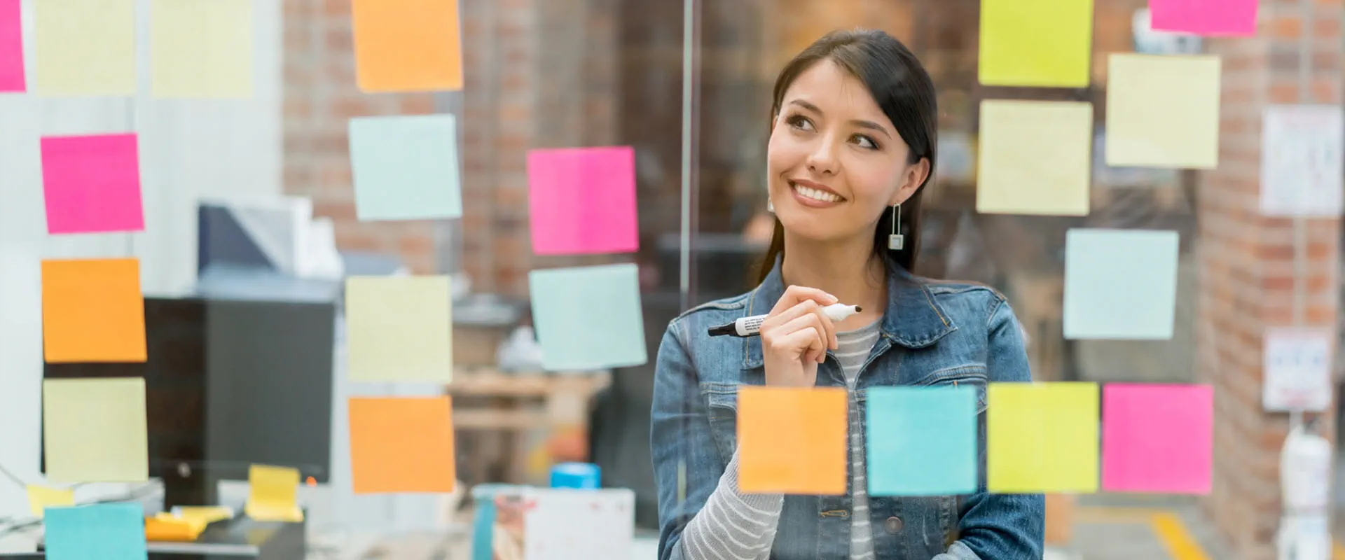 A casually dressed worker looks at a clear dry erase board with a marker in their hand. The board is covered in bright post-its