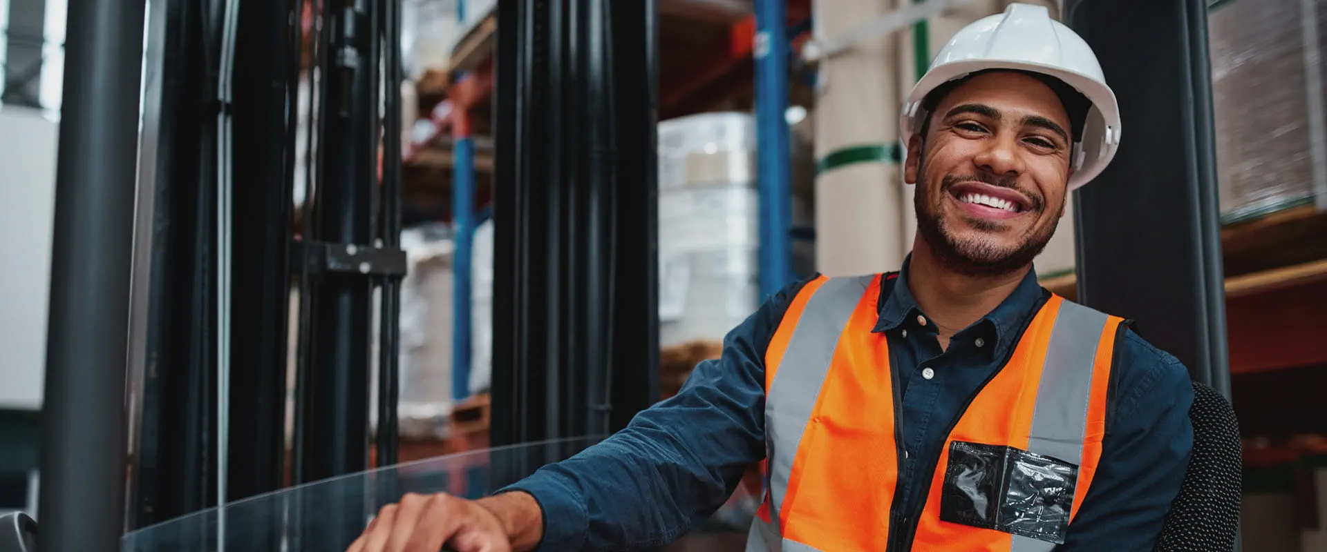  A smiling warehouse worker wearing a white hard hat, high-visibility orange safety vest.