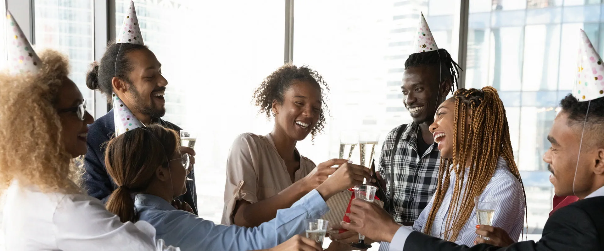 A diverse group of coworkers wearing party hats toast with drinks during an office celebration. 