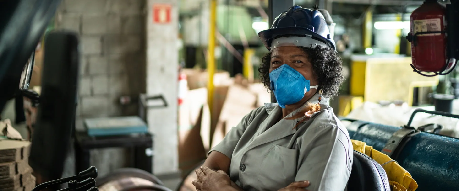 An older worker sits in a forklift, looking at the camera and wearing a mask