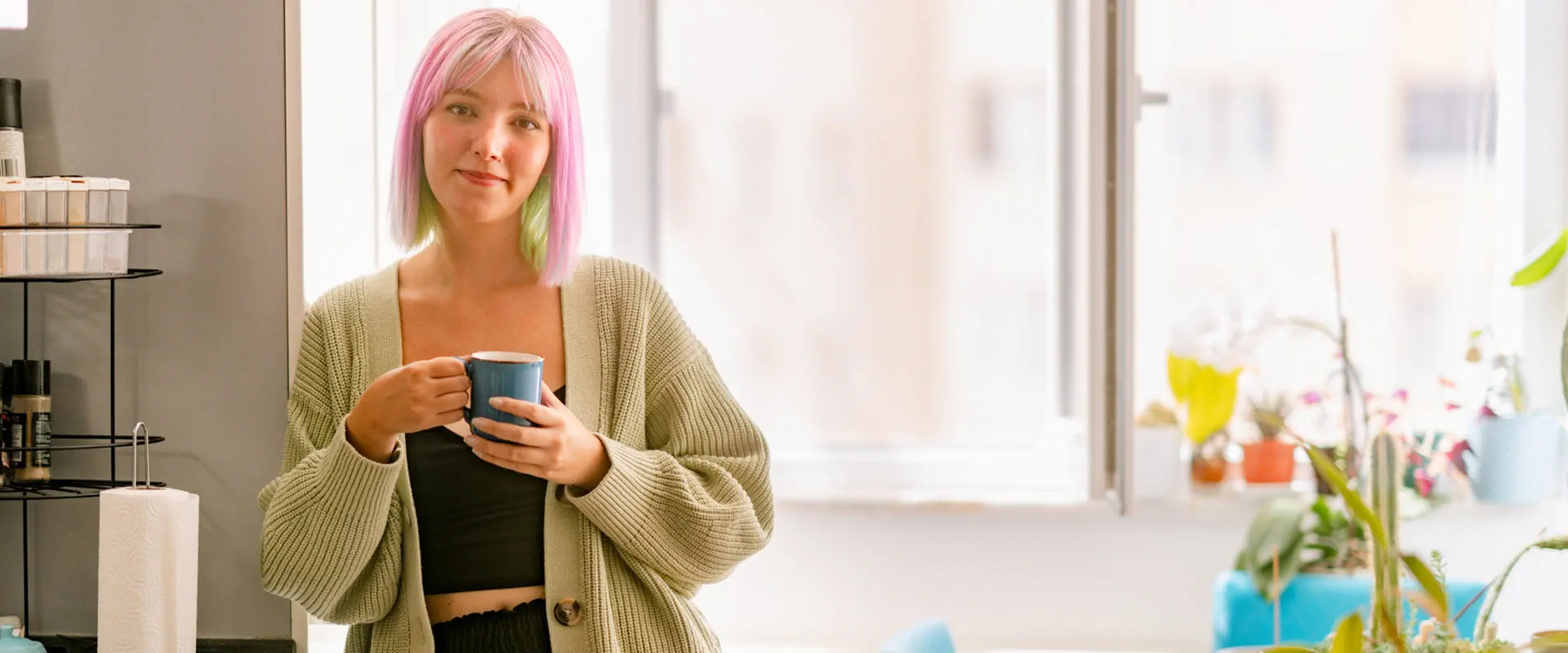 A young person, with pink and green dyed hair, in a crop top and cardigan leans against the wall and holds a blue mug, while smiling.