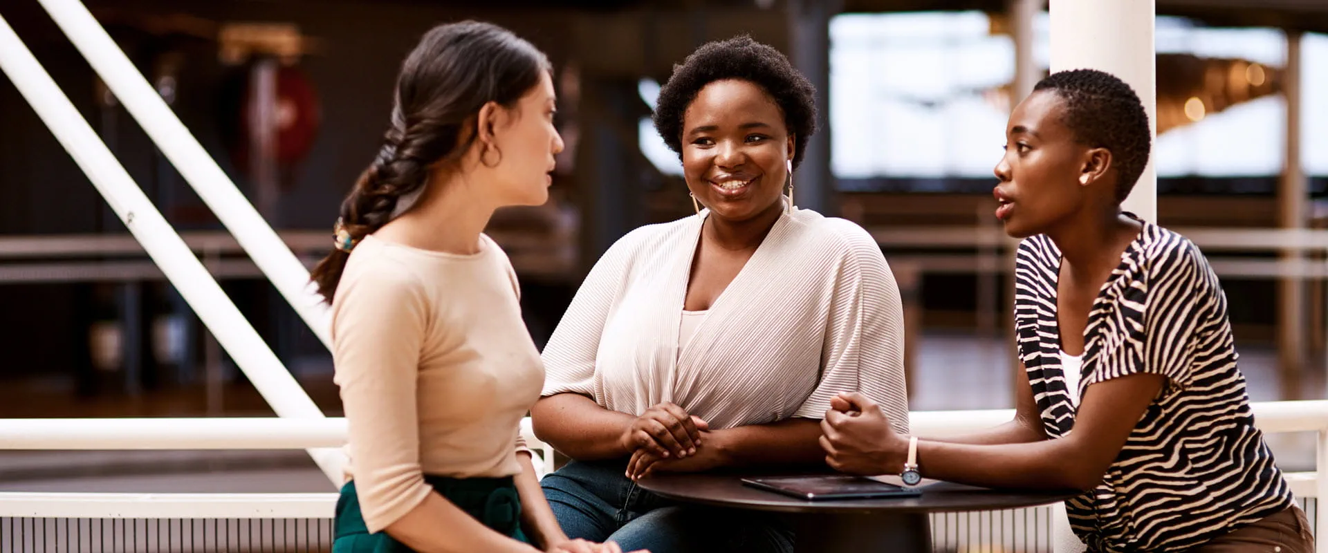 Three diverse individuals engaged in a friendly conversation at a modern office or coworking space. 