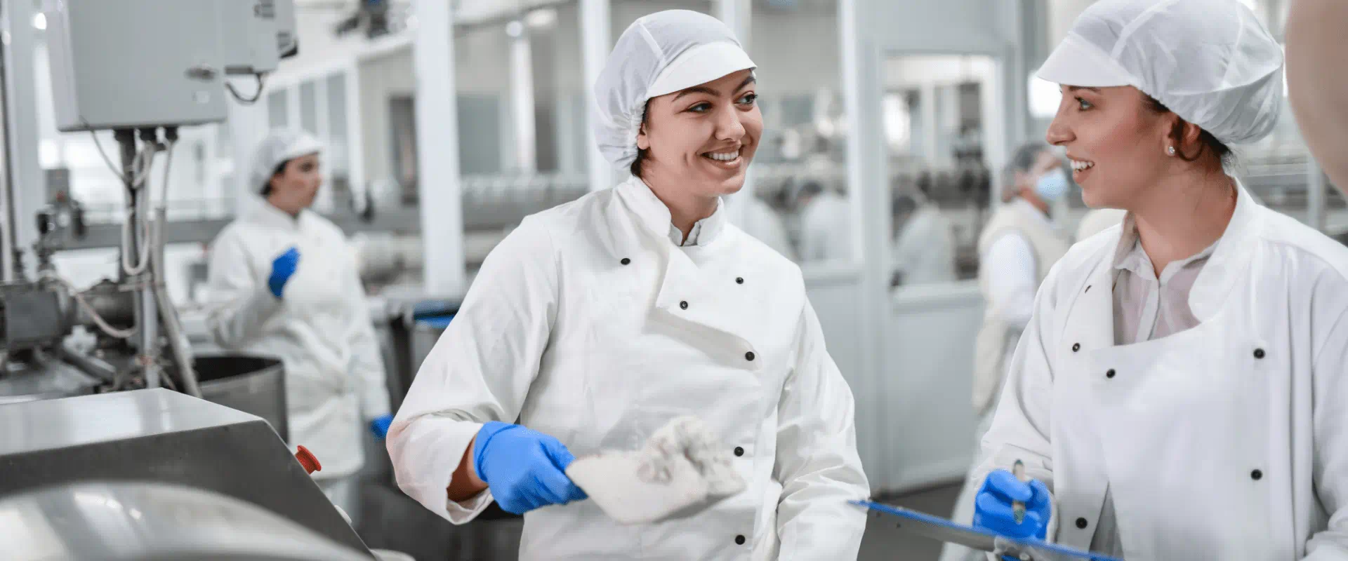 Two workers in a food processing plant are wearing hygienic clothing and hair coverings. They are laughing and talking.