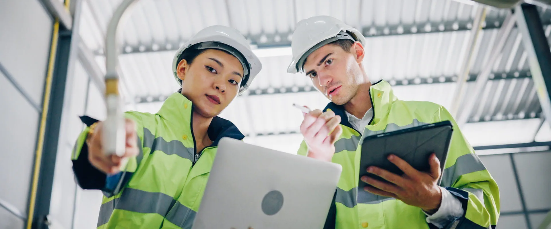 Two workers in safety jackets and hard hats look at a robotic arm in a manufacturing facility.