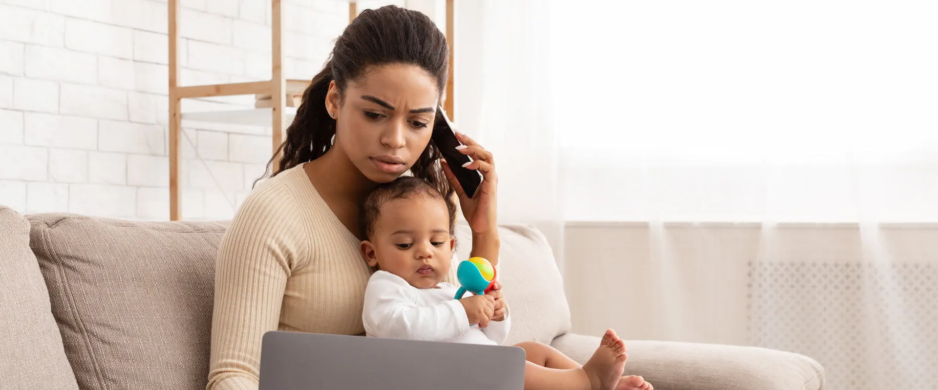 : A parent working from home sits on a couch with a laptop, holding a baby.