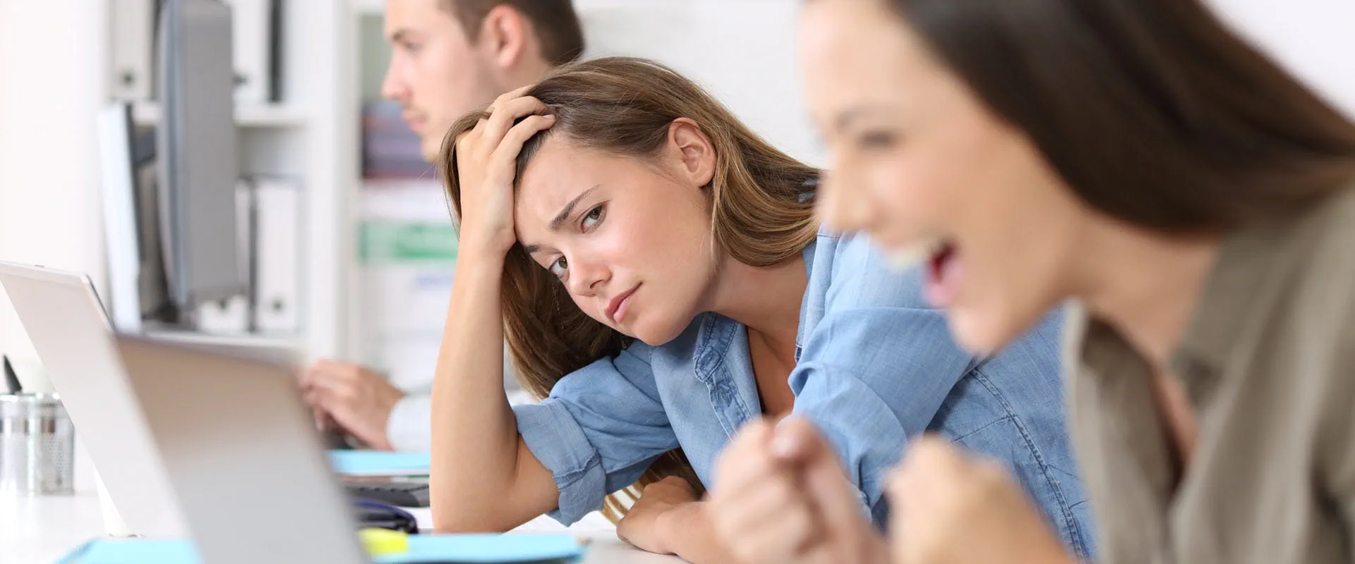 Three workers sit in a row working at laptops. The one in the middle looks annoyed at the person to her left, who is cheering with happiness