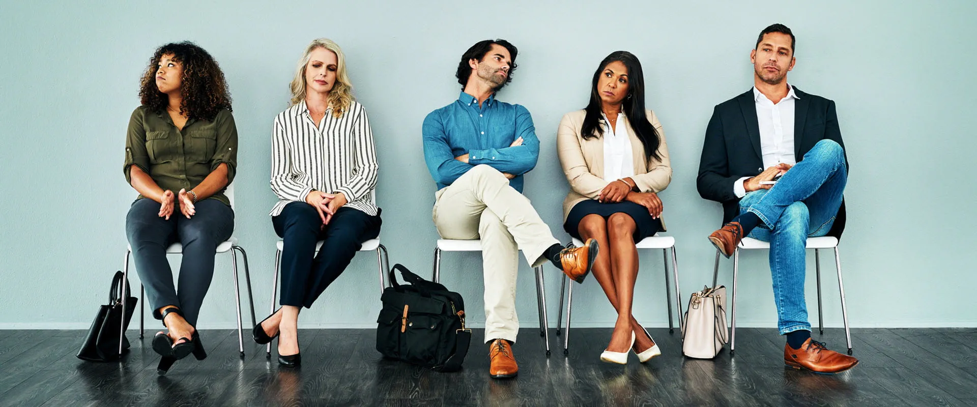Five workers in business casual dress sit on white plastic chairs, looking bored or irritated