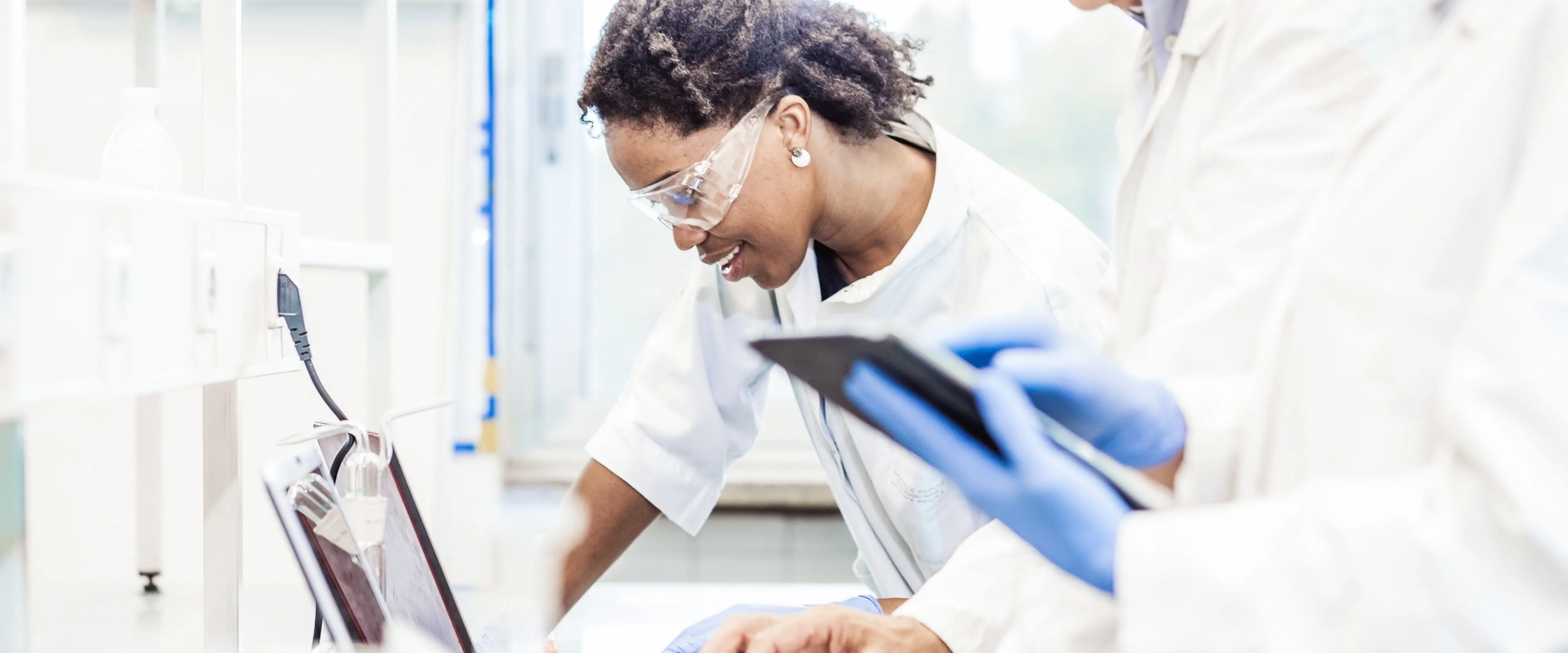 A worker in a white lab coat and safey glasses, leans over to type at a laptop. There are other workers in lab coats around them.
