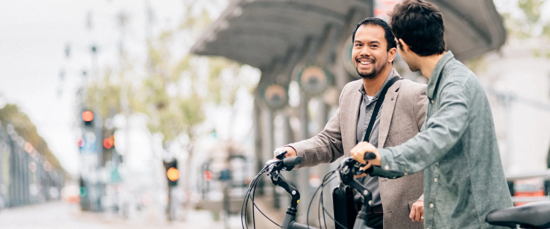 Two individuals in casual business dress talk to one another outside. Each is walking a bicycle.