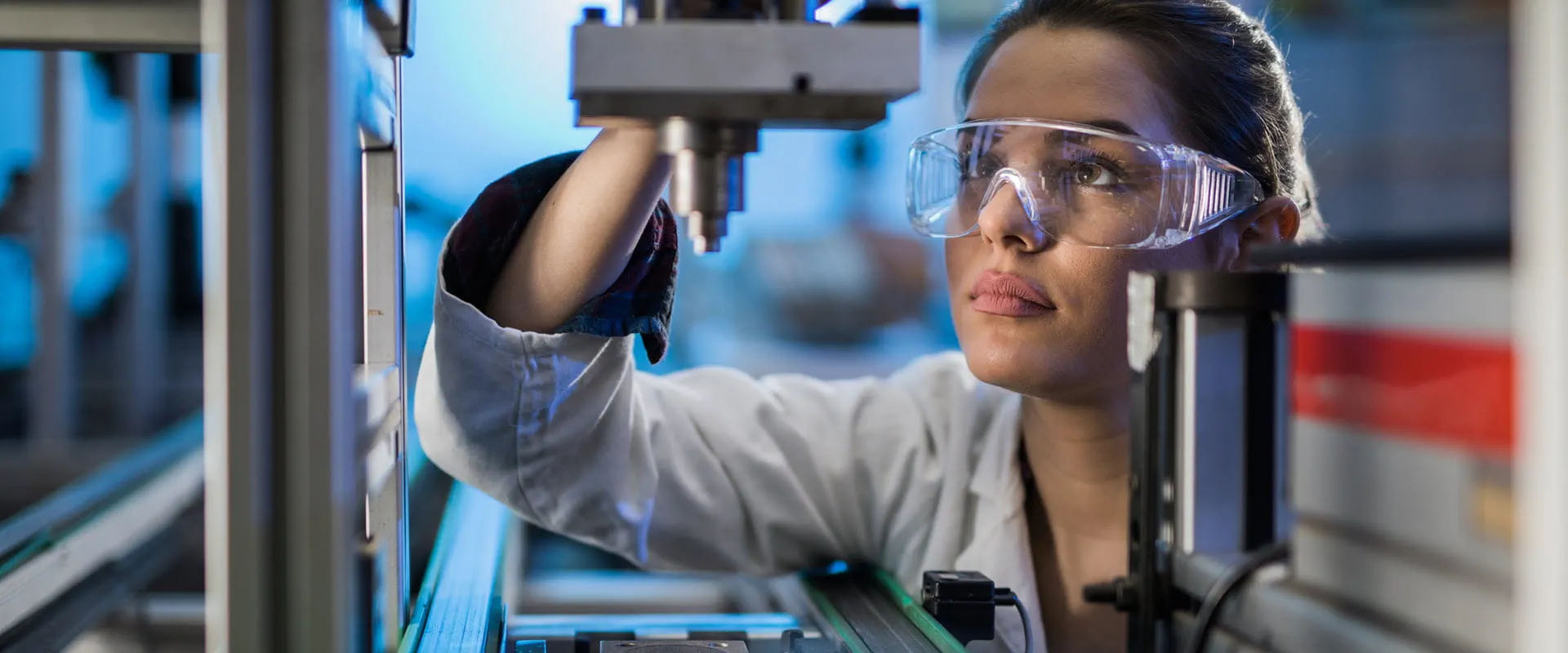 A professional wearing safety goggles and a lab coat closely inspecting a piece of industrial machinery in a high-tech manufacturing setting.